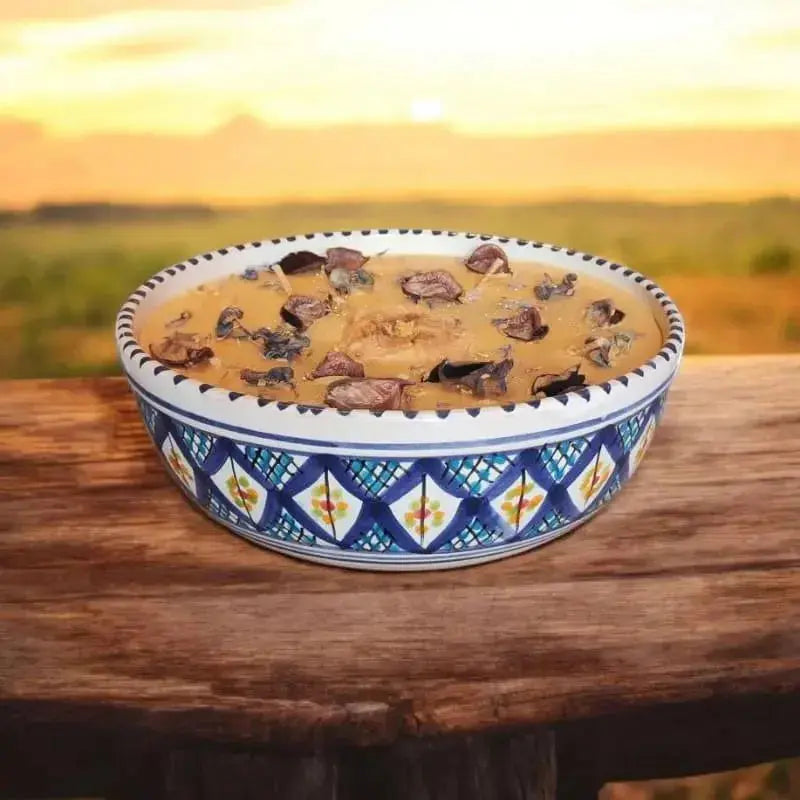 Bougie artisanale Lulu Baladine dans un bol bleu décoré de motifs géométriques, ornée de fleurs séchées, posée sur une table en bois face à un coucher de soleil.