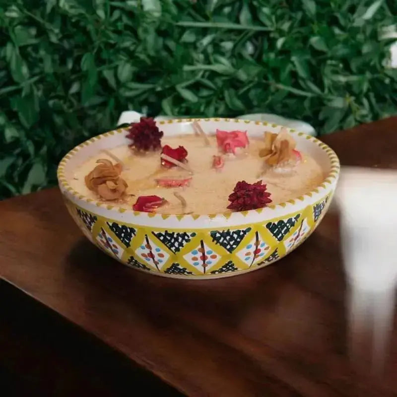 Bougie artisanale Lulu Baladine dans un bol jaune décoré de motifs, ornée de fleurs séchées rouges et roses, posée sur une table en bois.