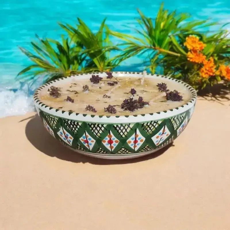 Bougie artisanale Lulu Baladine dans un bol vert décoré de motifs, ornée de fleurs séchées, posée sur le sable face à la mer turquoise.