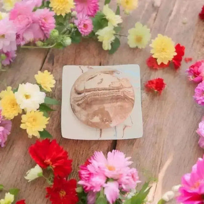 Une coupelle en argile carré avec un galet de massage de couleur beige clair et foncé entouré de fleur de couleur jaune, rose et rouge 